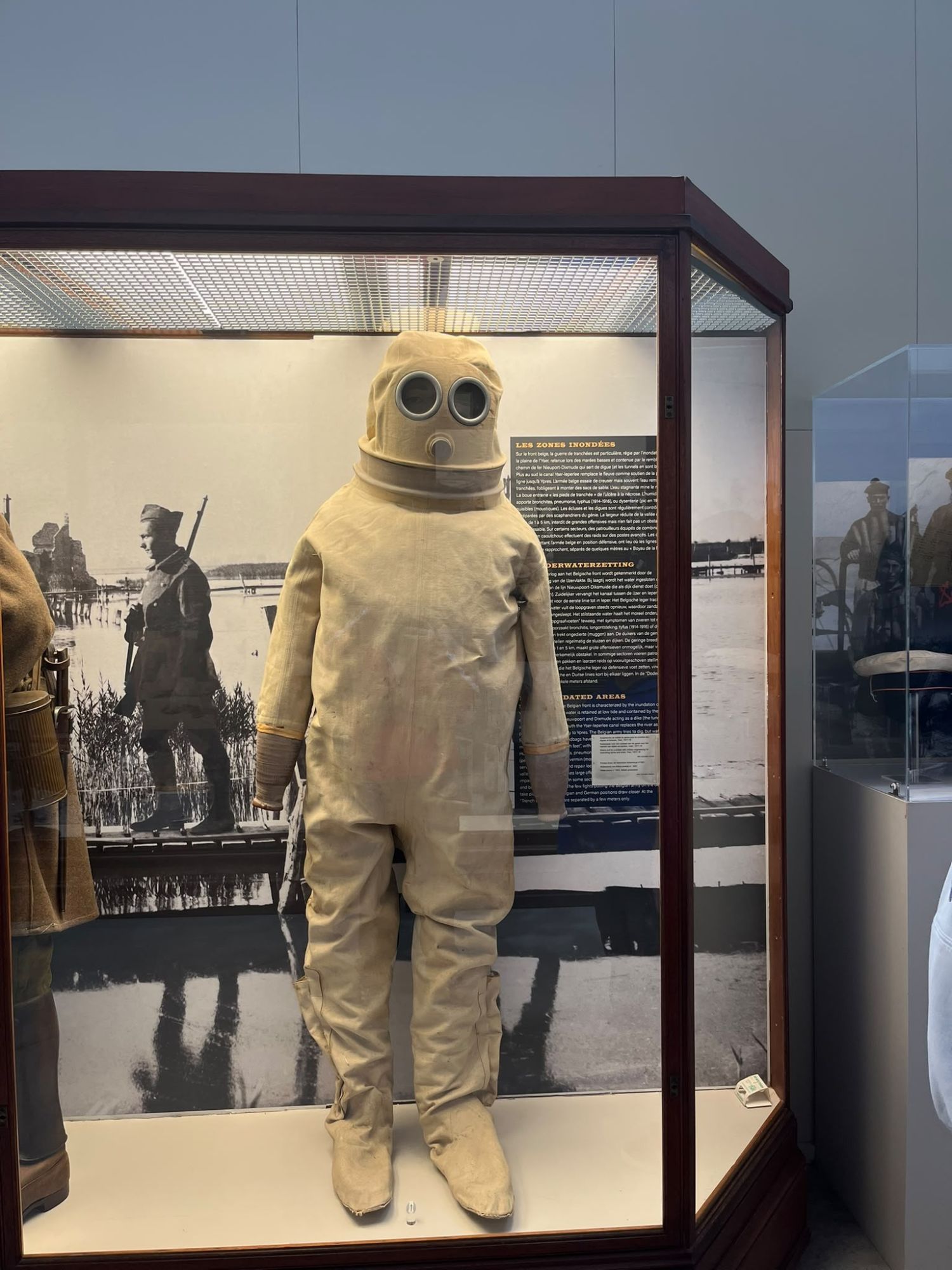A tan rubber protective suit with a round-lensed hood, displayed in a museum cabinet at the Brussels Air Museum, in front of a bilingual information panel about the flooded Yser front.